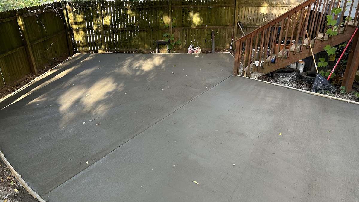 Freshly finished concrete patio with a curved edge beside a wooden deck and fenced backyard.